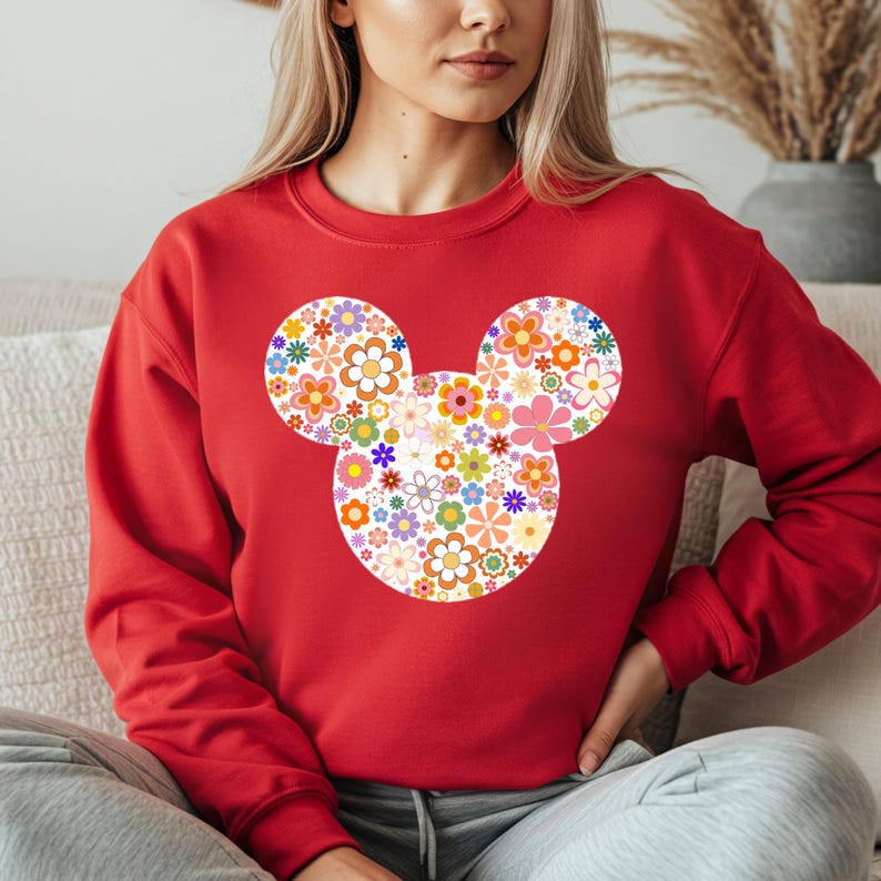 May include: A red sweatshirt featuring a floral Mickey Mouse design. The Mickey Mouse silhouette is filled with a variety of colorful flowers in shades of orange, pink, yellow, and purple. The sweatshirt is a solid red color.