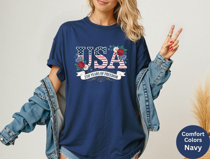 May include: Navy blue t-shirt with the word "USA" in red, white, and blue, decorated with flowers. The text "250 years of freedom" is displayed on a white banner. The shirt is part of the Comfort Colors collection.