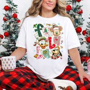 K&ouml;nnte beinhalten: Wei&szlig;es T-Shirt mit dem Wort "FALL" in bunten, festlichen Buchstaben. Jeder Buchstabe hat ein anderes Design, darunter Zuckerstangen, Mickey Mouse, Donald Duck und Pluto. Das Shirt ist mit Schneeflocken und Weihnachtsschmuck verziert.
