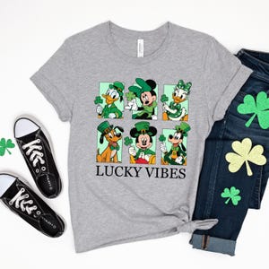 May include: A gray t-shirt with a graphic of Mickey Mouse, Minnie Mouse, Donald Duck, and Goofy dressed in green shamrock hats and holding shamrocks. The text "LUCKY VIBES" is printed below the graphic.
