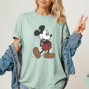 Puede incluir: Camiseta verde claro con un gr&aacute;fico de Mickey Mouse. El personaje de dibujos animados se muestra en negro, rojo y marr&oacute;n. La camiseta forma parte de la colecci&oacute;n Comfort Colors Bay.