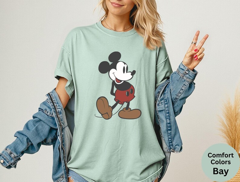Puede incluir: Camiseta verde claro con un gr&aacute;fico de Mickey Mouse. El personaje de dibujos animados se muestra en negro, rojo y marr&oacute;n. La camiseta forma parte de la colecci&oacute;n Comfort Colors Bay.