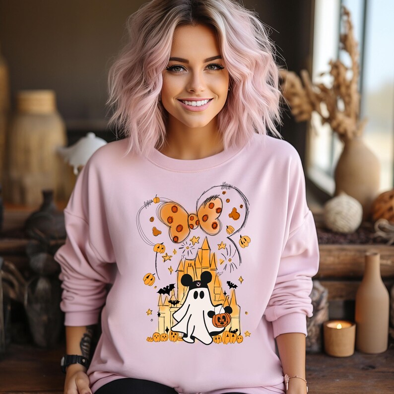 May include: Pink sweatshirt with a graphic of a ghost wearing a Mickey Mouse hat and holding a pumpkin. The ghost is in front of a castle with fireworks and a bow with polka dots.