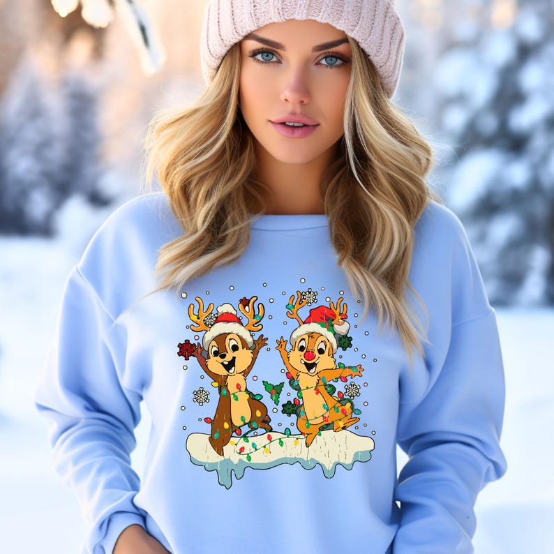 May include: A light blue sweatshirt featuring a Christmas design with two chipmunks dressed as reindeer with Christmas lights. The chipmunks are standing on a snowy surface with snowflakes falling around them.