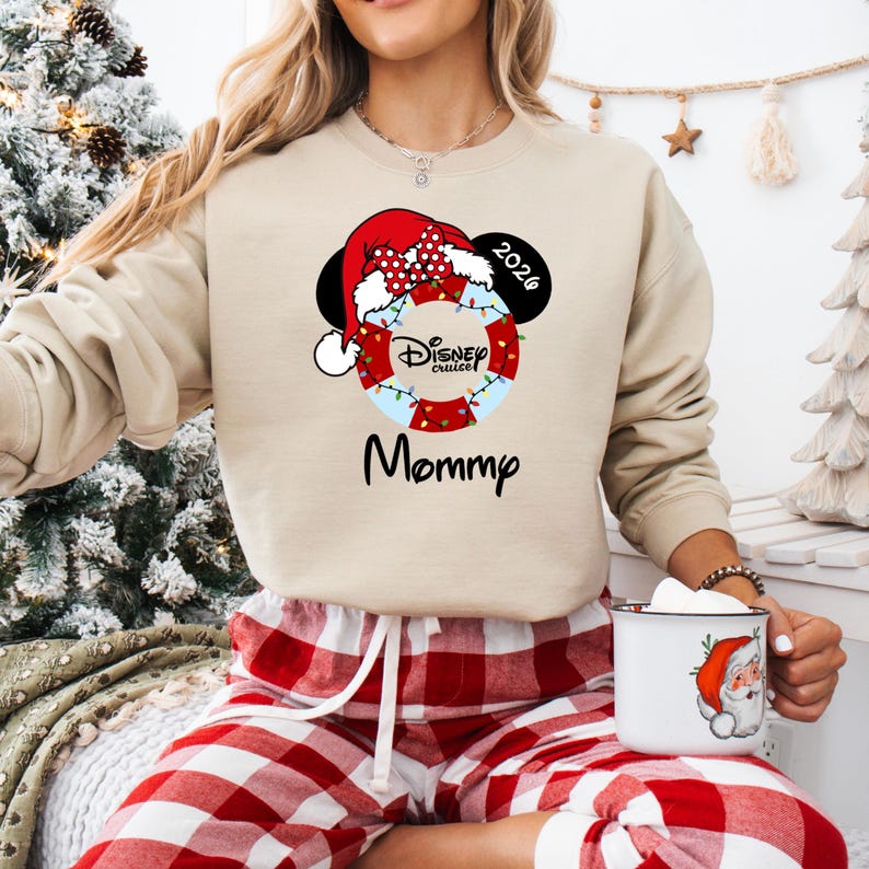 K&ouml;nnte beinhalten: Beiges Sweatshirt mit einem Disney Cruise-Design. Das Design umfasst einen Weihnachtsmannhut, Mickey Mouse-Ohren und die Texte "Mommp" und "2026". Das Design umfasst eine rot-wei&szlig; gepunktete Schleife und eine Rettungsboje.