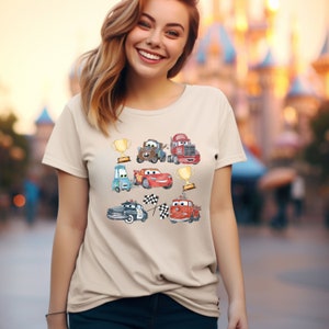 May include: A beige t-shirt with a colorful cartoon design featuring various cars from the Disney Pixar movie Cars. The design includes Lightning McQueen, Mater, and other characters from the film.