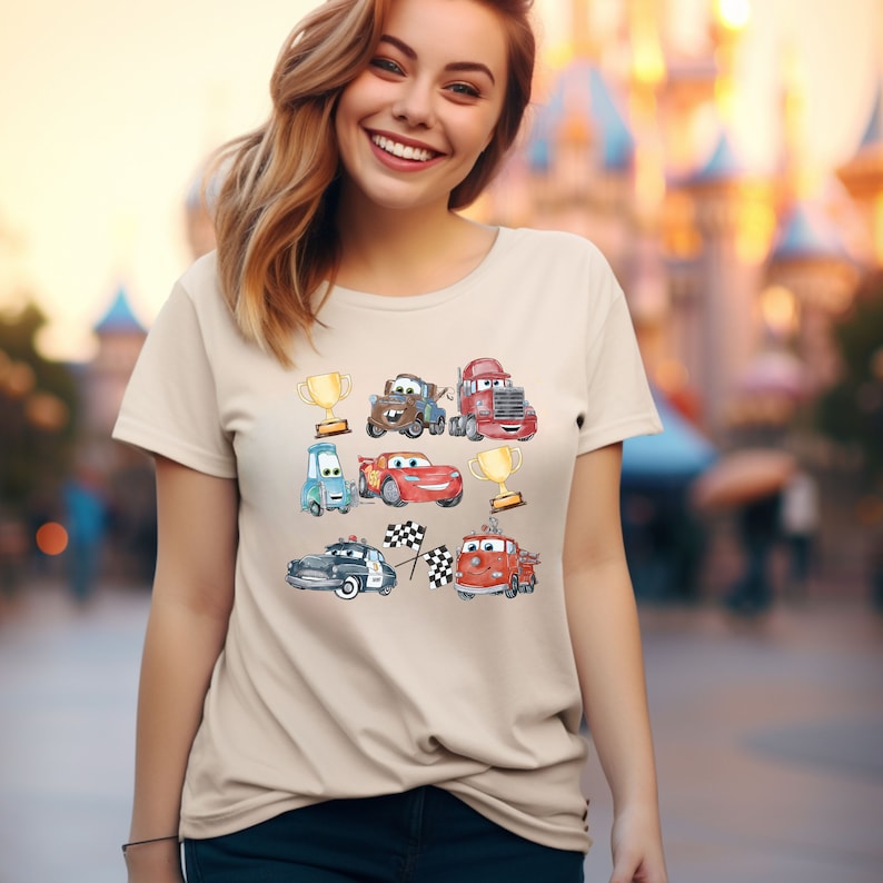 May include: A beige t-shirt with a colorful cartoon design featuring various cars from the Disney Pixar movie Cars. The design includes Lightning McQueen, Mater, and other characters from the film.