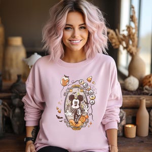 May include: A light pink sweatshirt featuring a Halloween-themed graphic. The graphic includes a skeleton Mickey Mouse in a decorative frame, surrounded by candy corn, bats, and the word "Spooky". The sweatshirt has long sleeves.