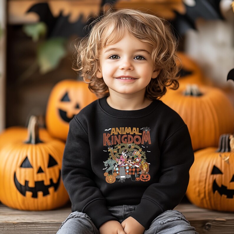Op de afbeelding: Een kind draagt een zwarte trui met de woorden "ANIMAL KINGDOM" in oranje en bruin. De trui heeft stripfiguren en pompoenen. De achtergrond bevat uitgesneden pompoenen en Halloween-decoraties.