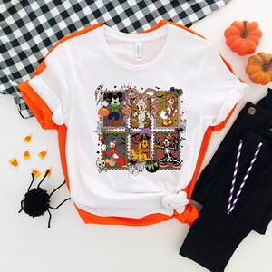 以下が含まれることがあります： ハロウィーンをテーマにしたカラフルなグラフィックが特徴の白いTシャツ。コスチュームを着た漫画のキャラクターが描かれています。グラフィックには、ミッキーマウス、ミニーマウス、ドナルドダックなどが含まれています。オレンジ色のトリムと黒いパンツで折りたたまれています。