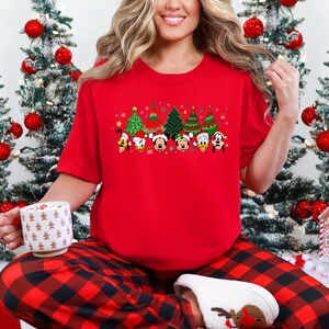 May include: A red short-sleeve t-shirt featuring a festive Christmas design. The design includes cartoon characters wearing Santa hats and Christmas trees. The person is wearing red and black plaid pajama pants and white slippers.