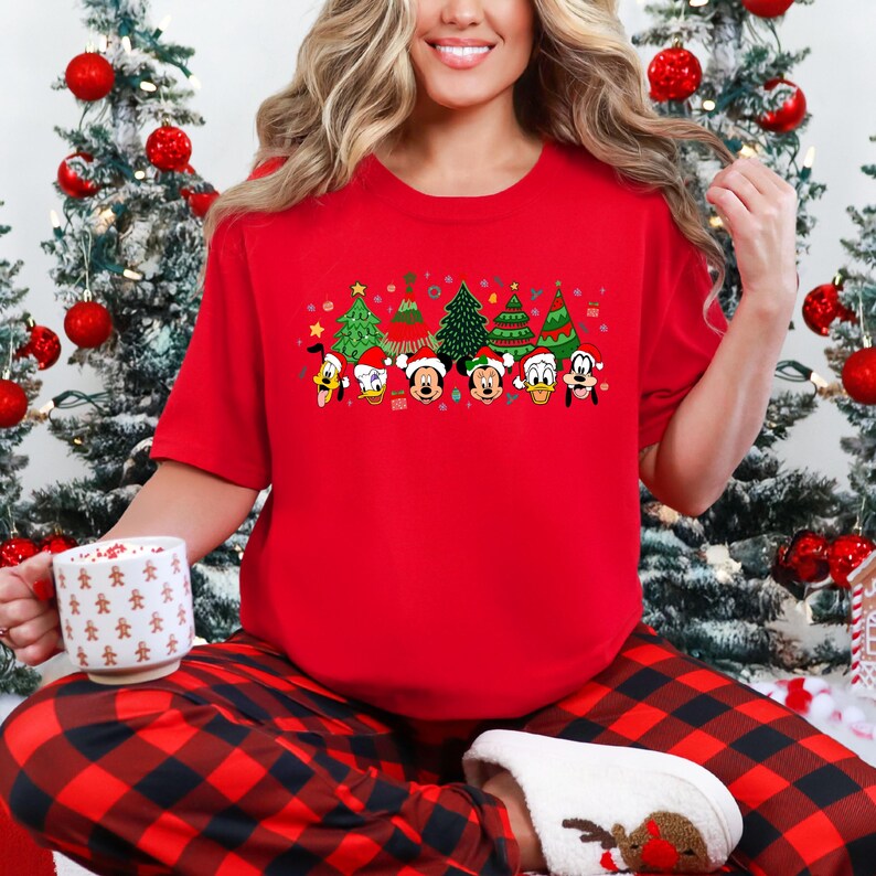 May include: A red short-sleeve t-shirt featuring a festive Christmas design. The design includes cartoon characters wearing Santa hats and Christmas trees. The person is wearing red and black plaid pajama pants and white slippers.