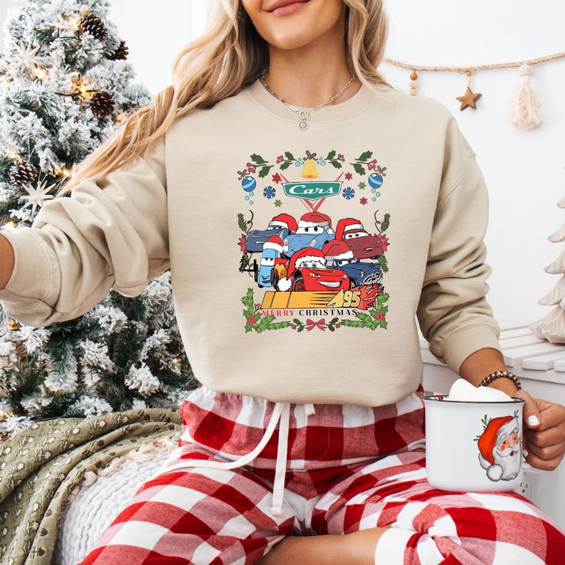 May include: Beige sweatshirt featuring a Cars-themed Christmas design. The graphic includes cartoon cars wearing Santa hats, the word "Cars", and "Merry Christmas" text. The sweatshirt is paired with red and white plaid pajama pants.