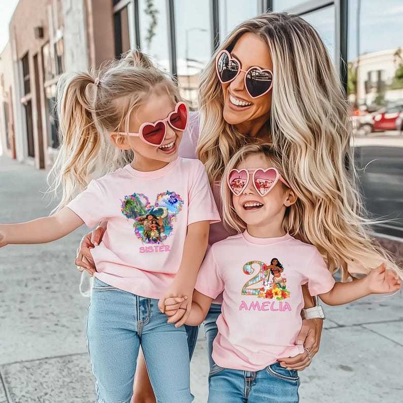 May include: Three people wearing pink t-shirts and heart-shaped sunglasses pose for a photo. The shirts feature colorful graphics and the names "Sister" and "Amelia". The group is smiling and holding hands, with a sunny outdoor background.