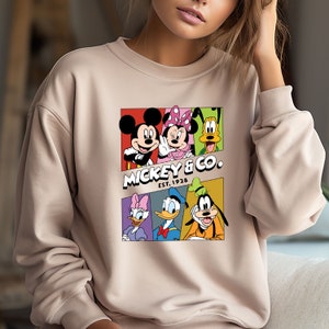 Pu&ograve; includere: Una felpa beige con una stampa grafica colorata di Topolino e i suoi amici. La grafica presenta Topolino, Minnie Mouse, Paperino, Pippo e Pluto in quadrati colorati. Il testo "Mickey & Co. Est. 1928" &egrave; stampato sotto la grafica.