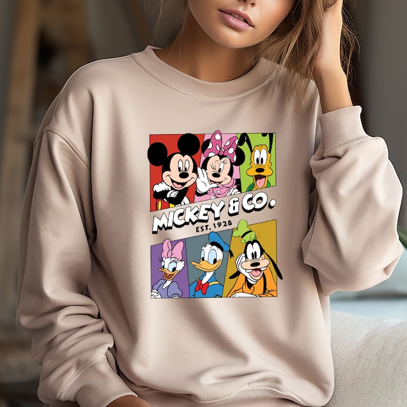 Pu&ograve; includere: Una felpa beige con una stampa grafica colorata di Topolino e i suoi amici. La grafica presenta Topolino, Minnie Mouse, Paperino, Pippo e Pluto in quadrati colorati. Il testo "Mickey & Co. Est. 1928" &egrave; stampato sotto la grafica.