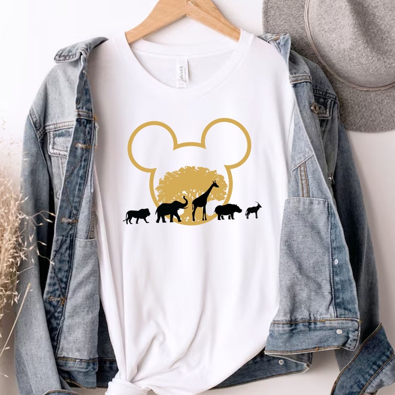 May include: White t-shirt with a gold Mickey Mouse head silhouette. Inside the silhouette is a brown tree with a giraffe, elephant, lion, hippopotamus, and antelope.