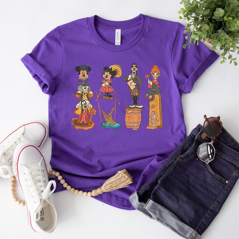 May include: Purple t-shirt featuring a graphic print of Mickey Mouse, Minnie Mouse, Donald Duck, Goofy, and a crocodile. The characters are all wearing different outfits and standing on barrels.