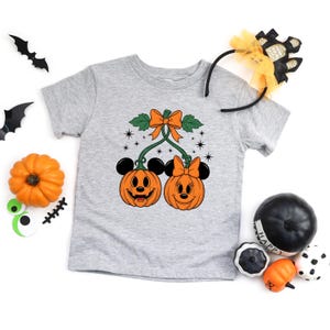 May include: A light gray t-shirt featuring a Halloween-themed graphic. The graphic includes two smiling pumpkin faces with mouse ears and bows, connected by a stem and bow. Additional Halloween decorations are scattered around the shirt.