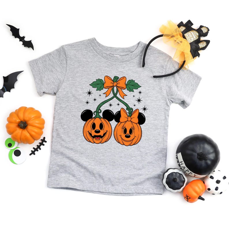 May include: A light gray t-shirt featuring a Halloween-themed graphic. The graphic includes two smiling pumpkin faces with mouse ears and bows, connected by a stem and bow. Additional Halloween decorations are scattered around the shirt.