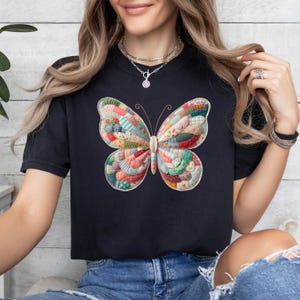 May include: Black t-shirt featuring a colorful butterfly design. The butterfly has patchwork-style wings with various fabric patterns in shades of red, green, blue, and yellow. The shirt is a casual, everyday wear item.