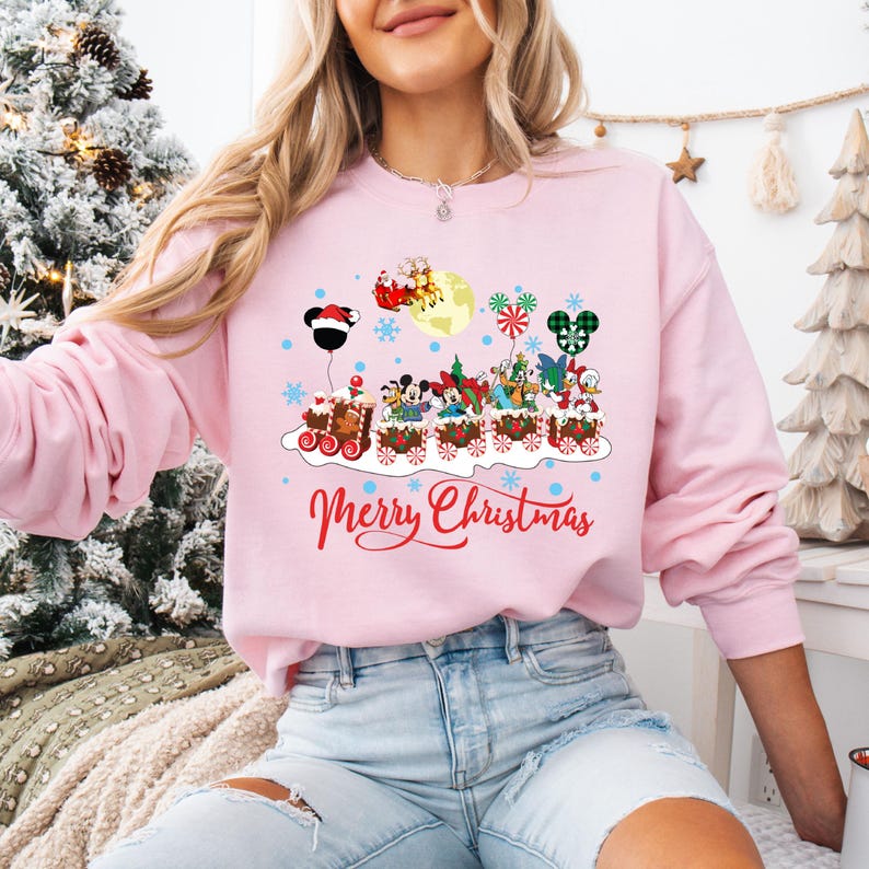 May include: Pink Christmas sweatshirt with a cartoon train design featuring Mickey Mouse and other Disney characters. The train is decorated with Christmas lights and ornaments. The text "Merry Christmas" is printed below the train.