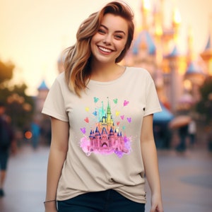 May include: A light beige t-shirt with a watercolor illustration of Cinderella's castle with rainbow colors and Mickey Mouse balloons.