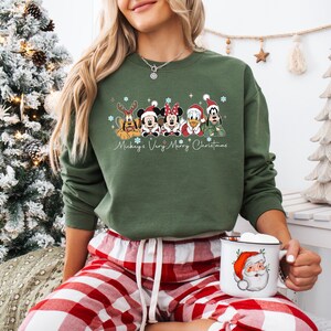 Puede incluir: Una sudadera verde oliva con un dise&ntilde;o navide&ntilde;o de Mickey, Minnie, Donald, Goofy y Pluto. El texto "Mickey's Very Merry Christmas" est&aacute; debajo. La persona lleva un pantal&oacute;n de pijama a cuadros rojos y blancos.