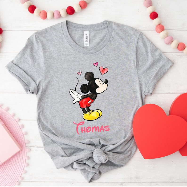 May include: A gray t-shirt featuring a cartoon Mickey Mouse blowing a kiss, with red hearts floating above. Mickey is wearing red shorts, yellow shoes, and white gloves. The name "Thomas" is written in pink script below.