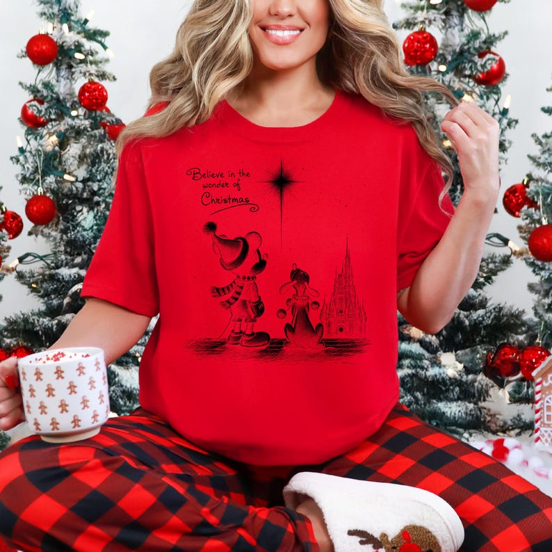 May include: A red t-shirt with a black graphic featuring Mickey Mouse and Pluto in Christmas attire, with a castle silhouette and the text "Believe in the wonder of Christmas." The person is wearing red and black plaid pajama pants.
