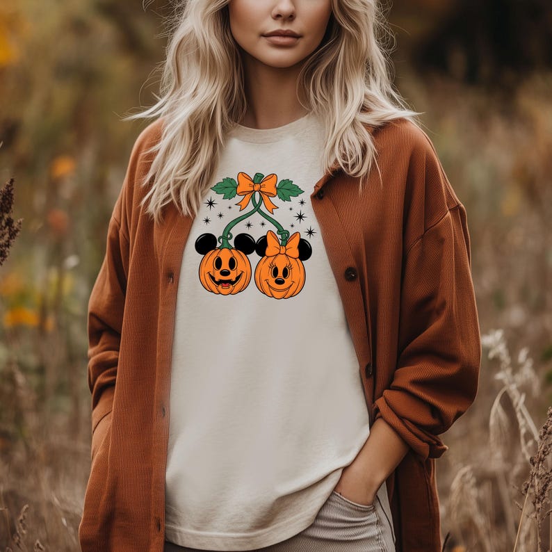 May include: A cream-colored t-shirt featuring two orange pumpkin faces with Mickey Mouse ears. The pumpkins are connected by green vines and a bow. The shirt is worn with a brown cardigan.