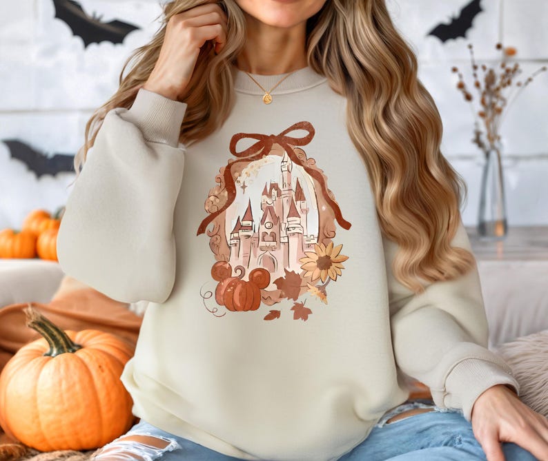 May include: Cream-coloured sweatshirt with a castle design, pumpkins, a sunflower, and autumn leaves. The design includes a brown ribbon and a Mickey Mouse pumpkin. The sweatshirt has long sleeves.