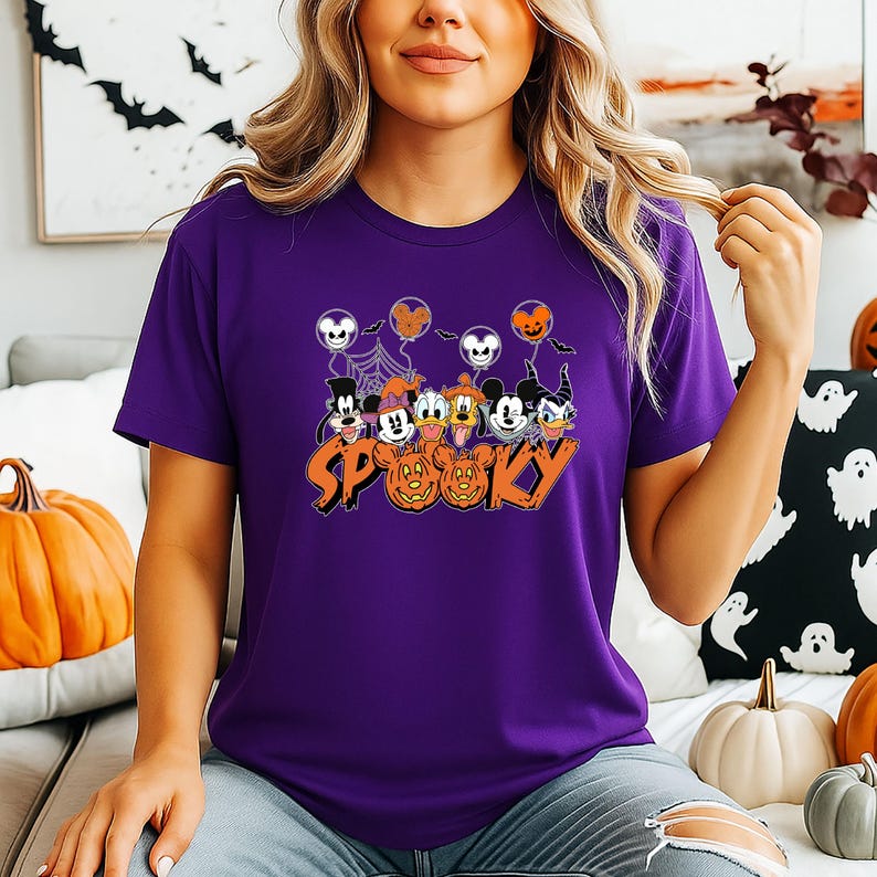 May include: A purple t-shirt with a Halloween design. The graphic features cartoon characters, balloons, pumpkins, and the word "Spooky" in orange. The t-shirt has short sleeves.