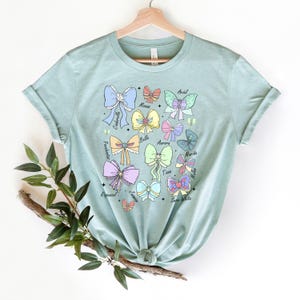May include: A light blue t-shirt with a graphic of bows and the names of Disney princesses: Ariel, Moana, Cinderella, Belle, Aurora, Merida, Mulan, Tiana, Rapunzel, Jasmine, Snow White.
