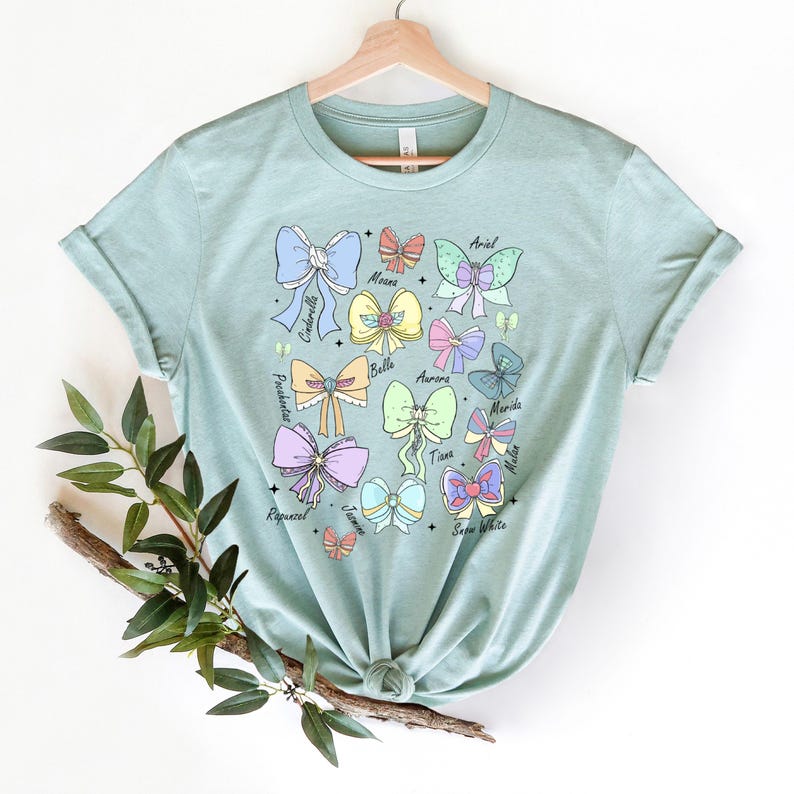 May include: A light blue t-shirt with a graphic of bows and the names of Disney princesses: Ariel, Moana, Cinderella, Belle, Aurora, Merida, Mulan, Tiana, Rapunzel, Jasmine, Snow White.