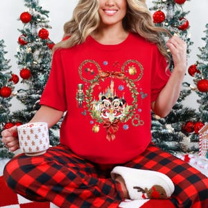 May include: A red short-sleeve shirt featuring a Christmas-themed Mickey and Minnie Mouse design. The shirt is paired with red and black plaid pajama pants. A white mug with gold stars is held in the left hand.