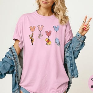 May include: Pink t-shirt featuring cartoon characters holding Mickey Mouse-shaped balloons. The characters include Tigger, Piglet, Winnie the Pooh, and Eeyore. The shirt has the words "Comfort Colors Blossom" on the bottom right.
