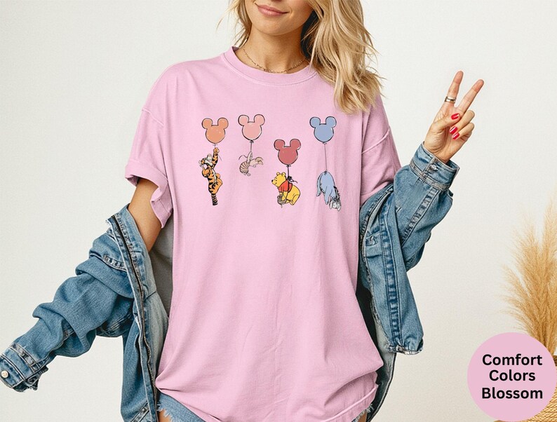 May include: Pink t-shirt featuring cartoon characters holding Mickey Mouse-shaped balloons. The characters include Tigger, Piglet, Winnie the Pooh, and Eeyore. The shirt has the words "Comfort Colors Blossom" on the bottom right.