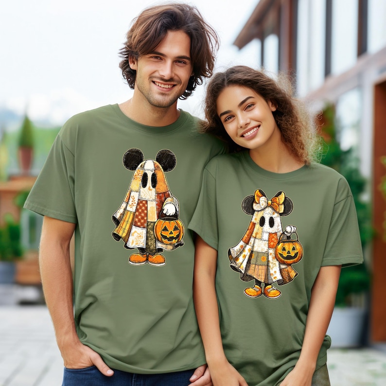 May include: Two olive green t-shirts featuring a cartoon ghost design with Mickey Mouse ears, a patchwork cloak, and a jack-o'-lantern. The shirts are worn by a man and a woman, perfect for Halloween.