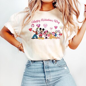 May include: A white t-shirt with a graphic of Mickey Mouse and friends holding heart-shaped balloons. The text "Happy Valentine's Day" is printed above the graphic.