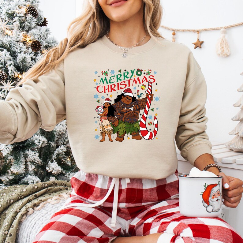 May include: Beige sweatshirt with a colorful graphic featuring the text "Merry Christmas" and cartoon characters. The design includes a candy cane and festive elements. The person is wearing red and white plaid pajama pants.