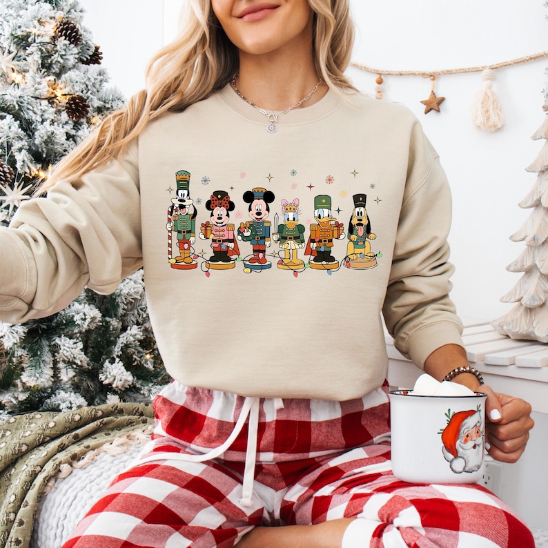 May include: Beige sweatshirt featuring a colorful graphic of Disney characters as nutcrackers. The characters include Mickey Mouse, Minnie Mouse, Donald Duck, and Goofy, each in festive outfits. The sweatshirt is paired with red and white plaid pajama pants.