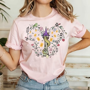 May include: Pink t-shirt with a floral design featuring a tower and a green chameleon. The design is in the shape of Mickey Mouse ears.