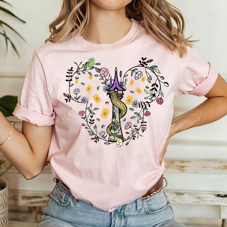 May include: Pink t-shirt with a floral design featuring a tower and a green chameleon. The design is in the shape of Mickey Mouse ears.