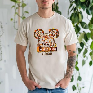 Puede incluir: Una camiseta beige con un dise&ntilde;o de orejas de Mickey Mouse hechas de hojas de oto&ntilde;o. El dise&ntilde;o incluye personajes de Disney y el texto "Thanksgiving Crew" en naranja y marr&oacute;n. La camiseta es de manga corta.