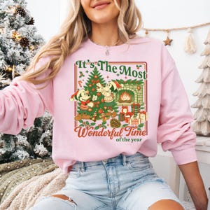May include: Pale pink sweatshirt with a Christmas-themed graphic. The design features Mickey and Minnie Mouse, a decorated tree, a fireplace, and the text "It's The Most Wonderful Time of the Year."