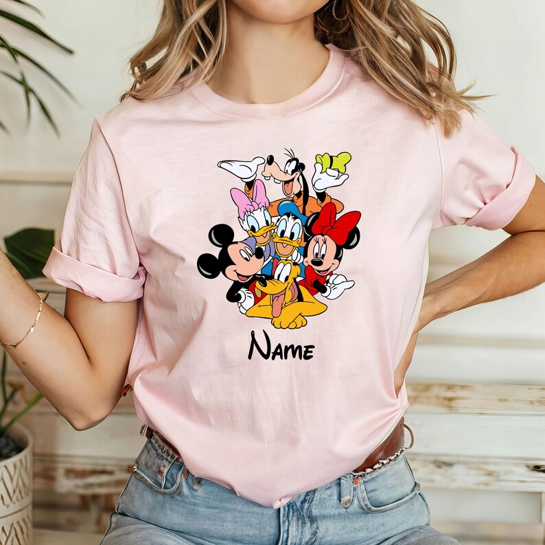 Op de afbeelding: Roze T-shirt met een afbeelding van Mickey Mouse, Minnie Mouse, Donald Duck, Goofy, Pluto en Daisy Duck. De tekst "NAME" is onder de afbeelding gedrukt.