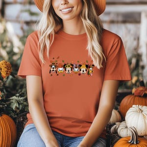 May include: An orange t-shirt featuring a graphic of Disney characters in fall attire. The graphic includes Mickey Mouse, Minnie Mouse, Donald Duck, Daisy Duck, Goofy, and Pluto, with autumn leaves and sunflowers. The shirt is a solid orange color.