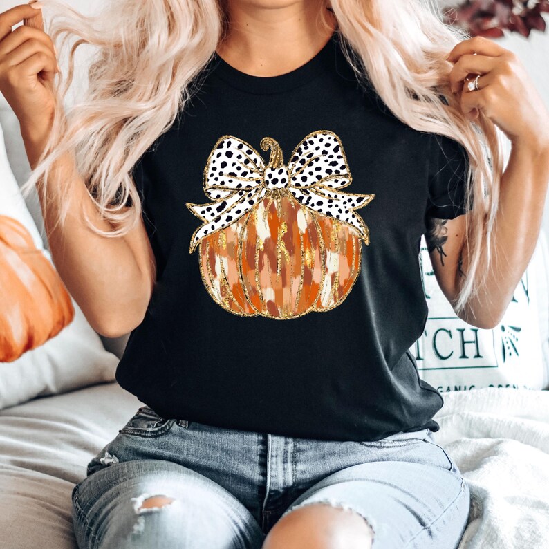 Puede incluir: Camiseta negra con un gr&aacute;fico de una calabaza con un lazo de lunares negros y blancos. La calabaza es naranja y marr&oacute;n con detalles de purpurina dorada.