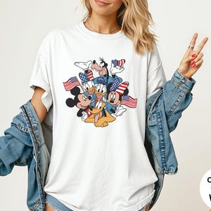 May include: White Comfort Colors t-shirt featuring a graphic of Mickey Mouse, Minnie Mouse, Donald Duck, Goofy, and Pluto, each holding or wearing patriotic American flags. The shirt has a crew neck and short sleeves.
